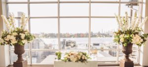 Two large floral arrangements frame a floral wedding centerpiece in front of a wall of windows.
