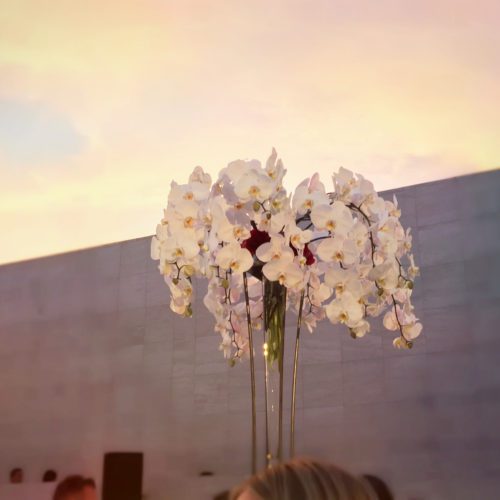 A huge white orchid arrangement in a tall glass vase is framed by the sunset at a corporate event.